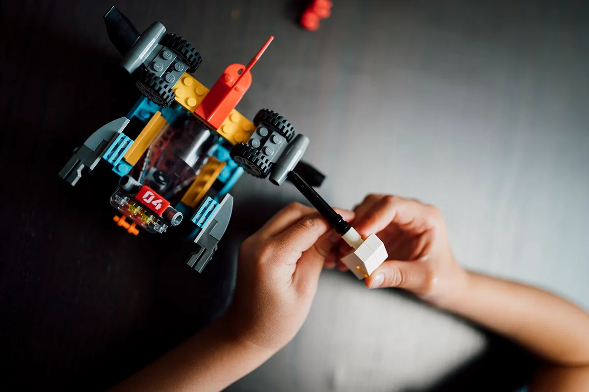 Kinderhanden bouwen aan een voertuig van gekleurde steentjes op een donkere tafel, met een los wit steentje tussen de vingers
